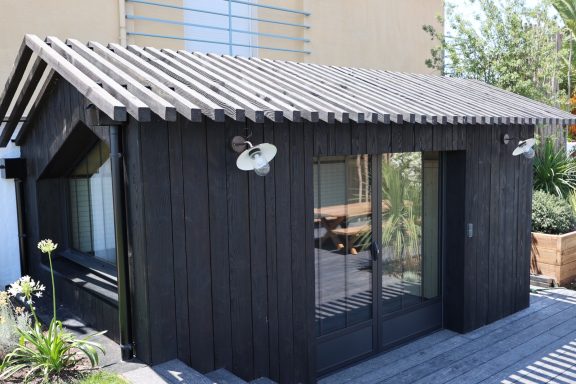 Façade en bois noir avec porte vitrée et applique extérieure sculpturale.