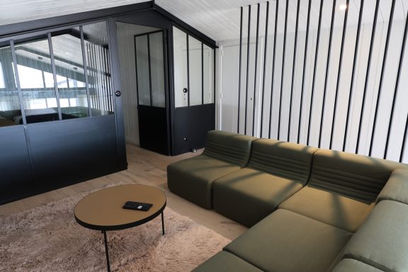 Salon secondaire avec canapé vert et verrière intérieure noire.